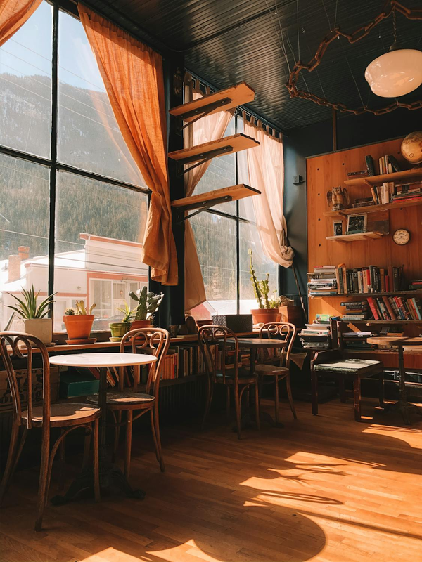 Café aconchegante com mesas e cadeiras de madeira dispostas perto de uma grande janela, convidando a um ambiente acolhedor e relaxante.